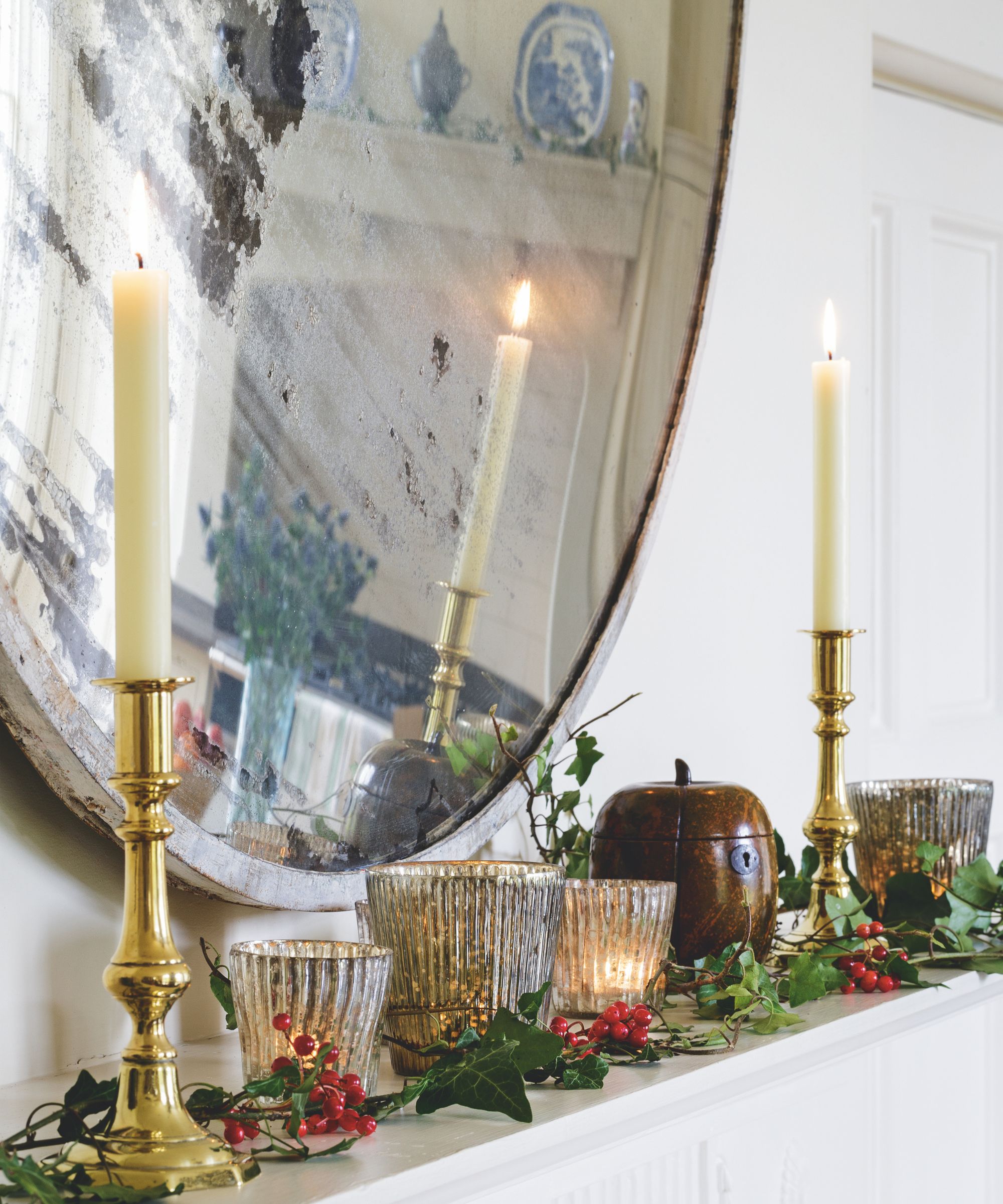 White taper candles in gold candle holders on a mantelpiece decorated with glass candle holders, greenery, red berries, and a large circular mirror above.