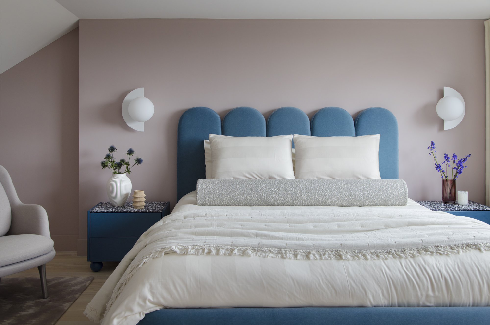 primary loft bedroom with pink walls, dark blue upholstered scalloped bedhead, blue bedside tables with vases of flowers, and armchair with wall sconces