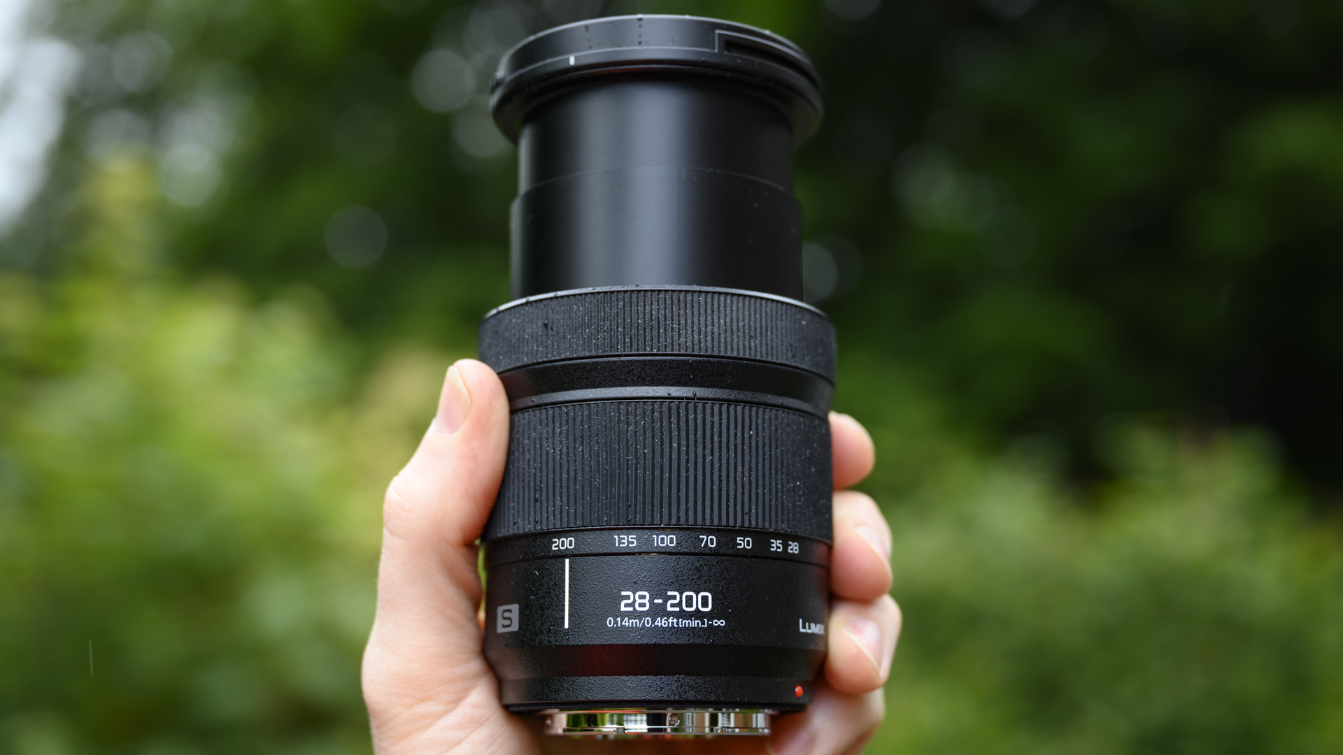 Panasonic Lumix S 28-200mm travel lens in the hand with leafy background and light rain