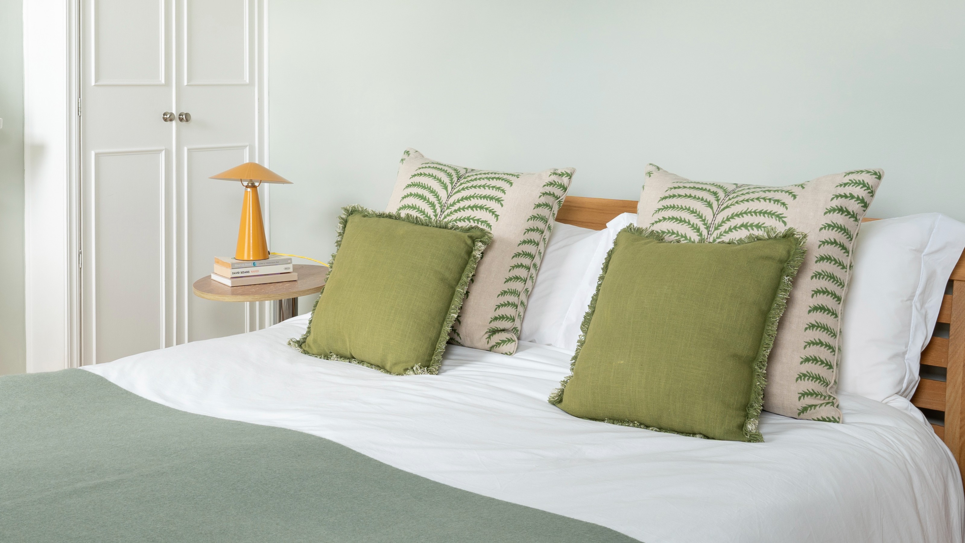 A soft green-painted bedroom with green cushions and throws on the bed and a yellow glossy metal table lamp from H&M on the bedside table