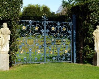Vintage garden gate in blue and gold wrought iron, flanked by hedging and two stone statues.