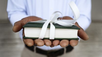 A man holds out a textbook wrapped in a white bow as a gift.