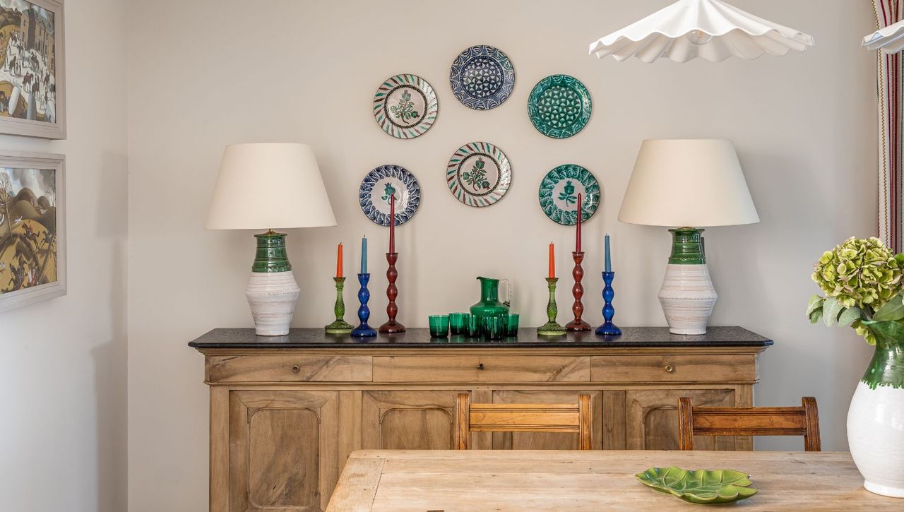 Corner of a white dining room with wooden sideboard, wooden dining table, plates on wall, table lamps, candles and flowers