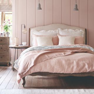 A pink bedroom with a wood panelled wall and a bed with a large upholstered headboard and ruffled pillows