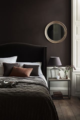 brown bedroom with brown upholstered bed, brown walls, round mirror, side table with glass based lamp and brown shade. brown and ochre blankets, cushions