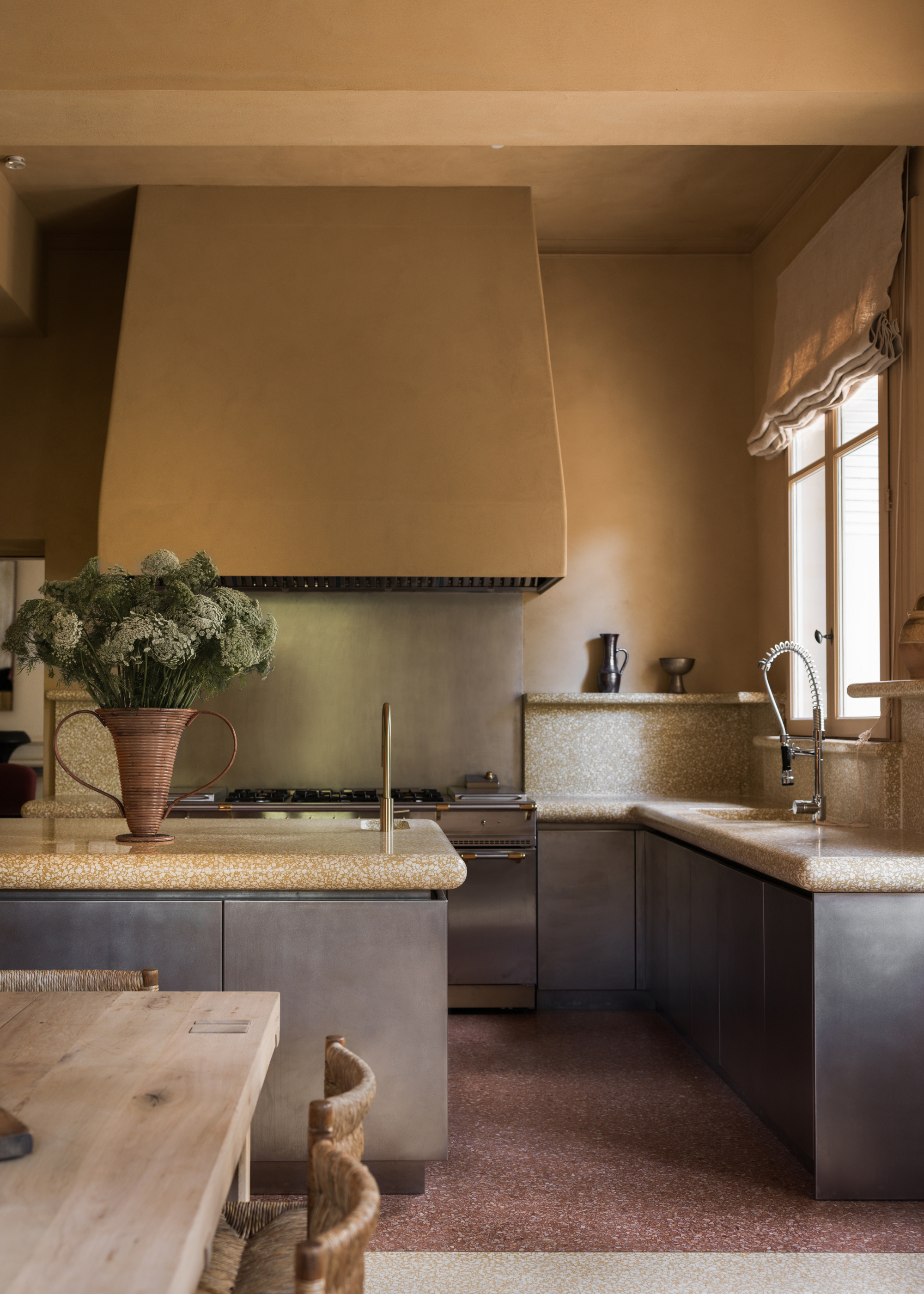 A kitchen with sandy walls, brushed steel cabinets, a large vase of flowers, and a wood dining table with woven chairs