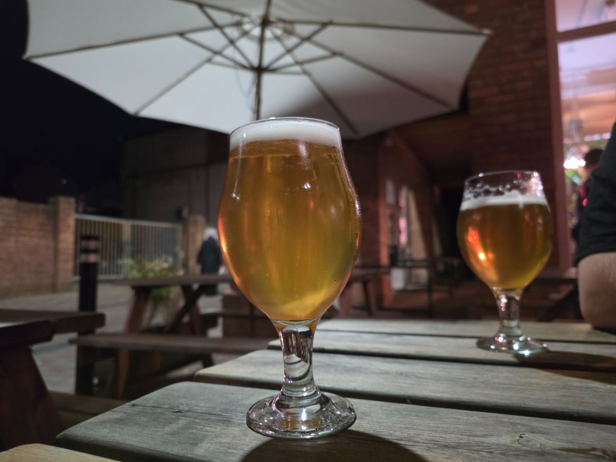 Two glasses of golden beer on a wooden outdoor pub table beneath a large patio umbrella.