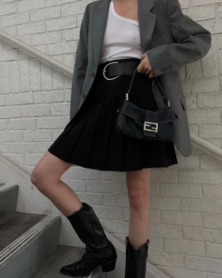 Woman standing on stairs wearing gray blazer, black skirt, Fendi baguette bag, and white tank top leaning on wall against stairwell.