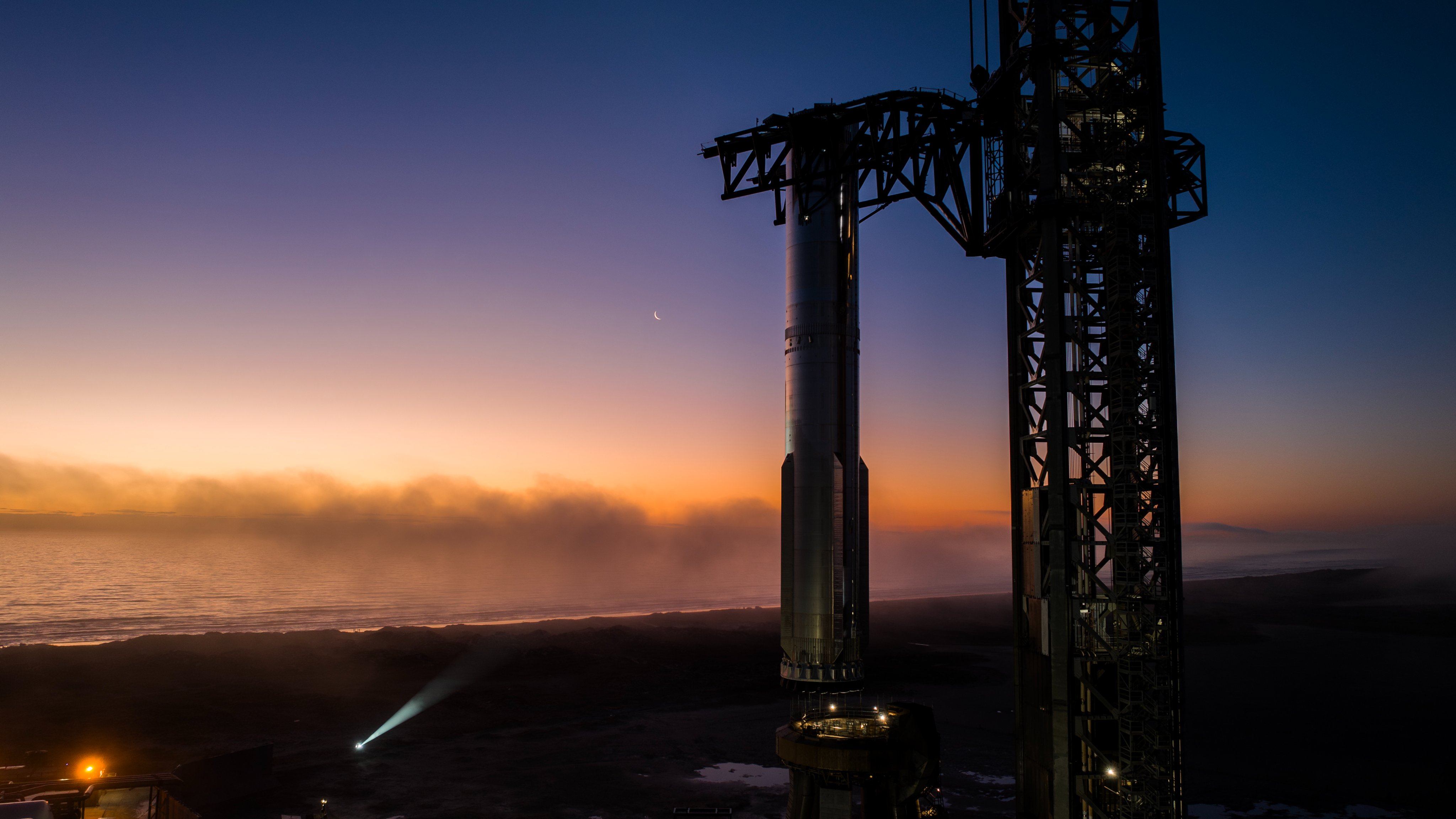 a silver rocket stands on a launch pad as dawn breaks on a foggy day