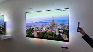 Philips OLED909 TV mounted on a white wall. On the screen is an aerial view across Barcelona.