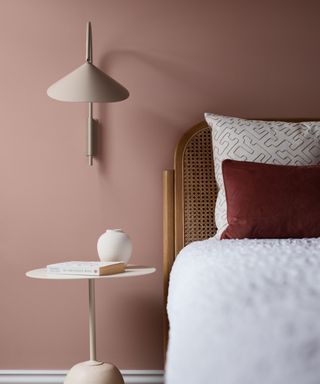 bedroom with earthy pink wall, rattan headboard, white bedding with dark red cushion and neutral minimal nightstand and wall sconce