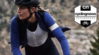 A white, female cyclist with blonde hair cycling head-on and close up to the camera, looking off to the side, wearing a blue Rapha jersey unzipped, to reveal a white Rapha base layer beneath, and a Cycling News 'Recommends' badge