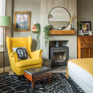 Grey living room with yellow armchair, wood burning stove, black rug, round mirror and bureau