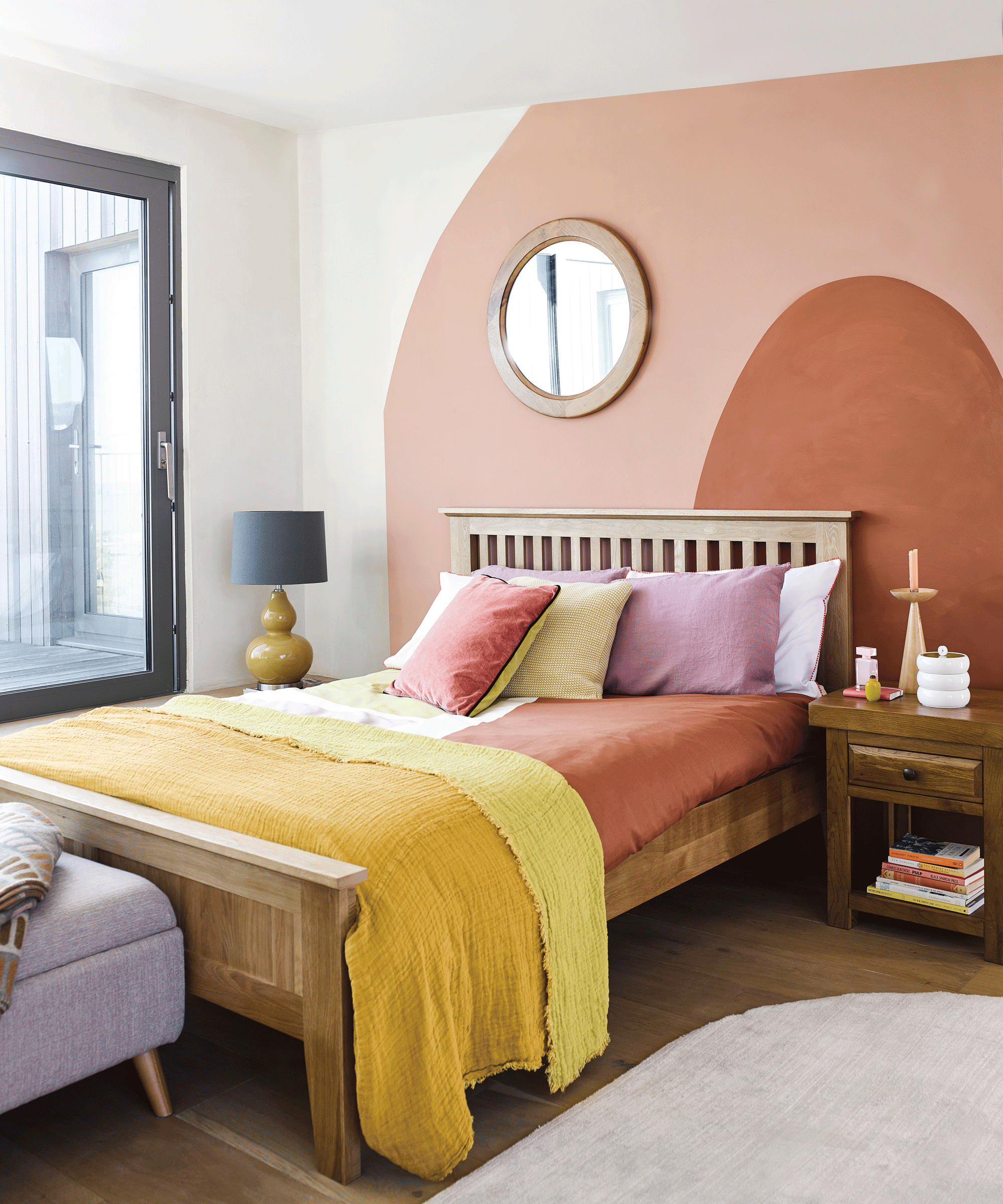 A bedroom with peach and terracotta feature wall with round framed mirror and yellow, rust and pink bedding
