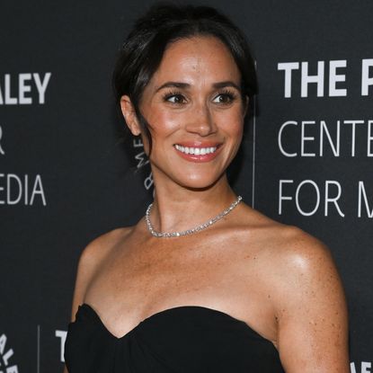 Meghan Markle in a black dress and diamond necklace posing in front of a gray backdrop with white lettering and smiling