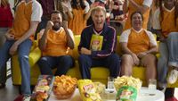 Actor Will Ferrell sits enthusiastically on a yellow sofa holding a bag of Lay's potato chips, flanked by two friends cheering and surrounded by bowls of chips, during a soccer-themed gathering.