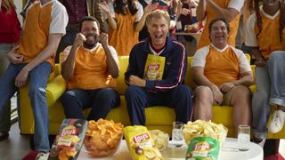 Actor Will Ferrell sits enthusiastically on a yellow sofa holding a bag of Lay's potato chips, flanked by two friends cheering and surrounded by bowls of chips, during a soccer-themed gathering.