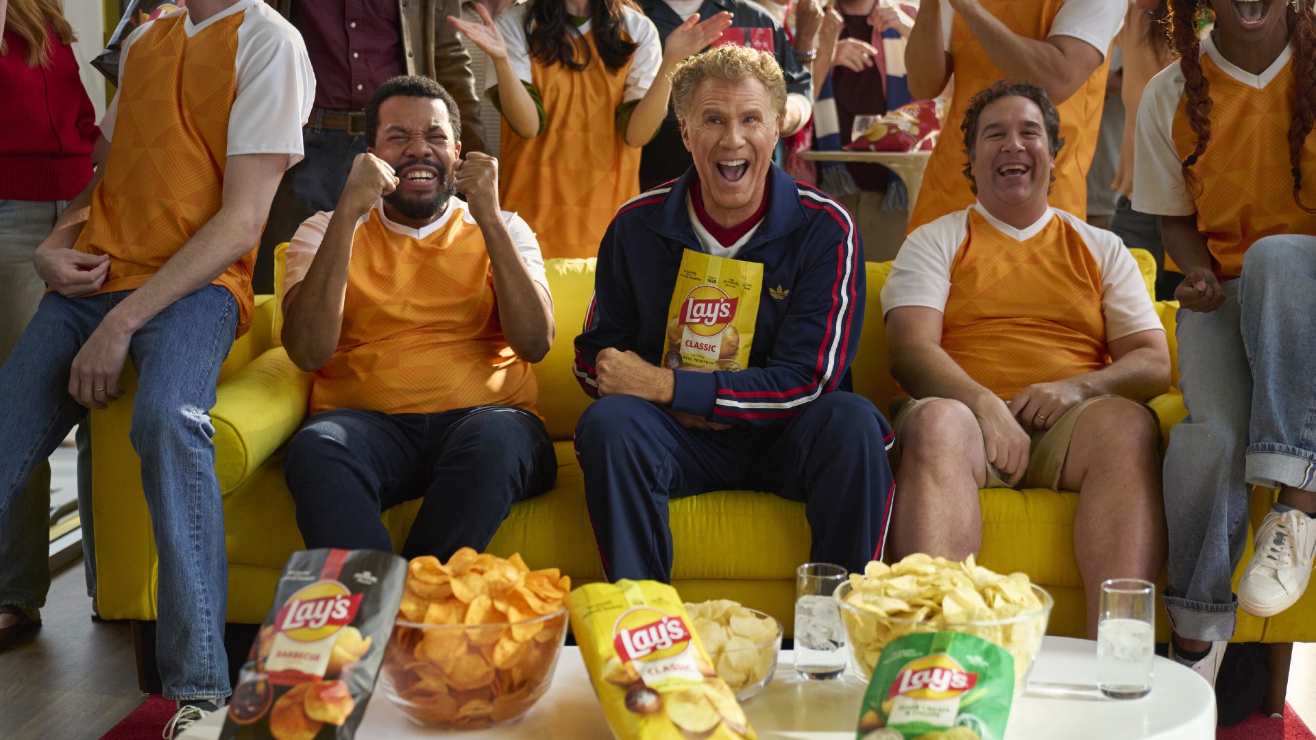 Actor Will Ferrell sits enthusiastically on a yellow sofa holding a bag of Lay's potato chips, flanked by two friends cheering and surrounded by bowls of chips, during a soccer-themed gathering.