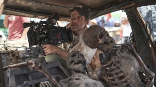 Zack Snyder holding camera in burned-out car set with two skeletons in front of him