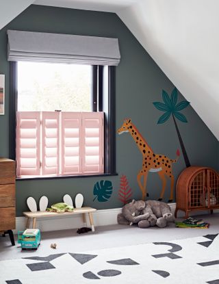 A playroom with sloped ceilings, pink shutters, gray walls and a giraffe mural.