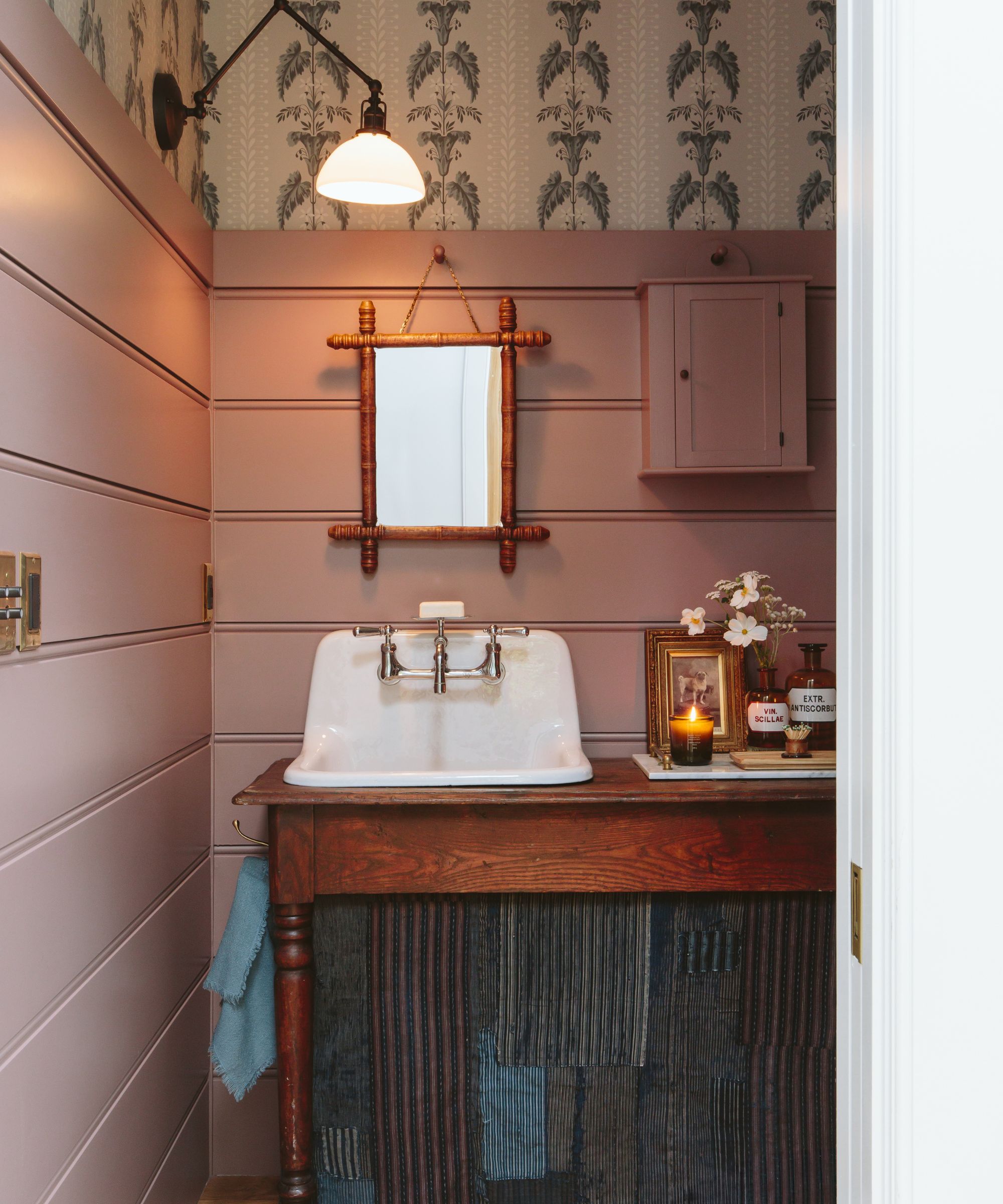 A powder room with mauve wall panelling, neutral wallpaper on the top portion of the wall, a wooden vanity unit, and rustic decor.