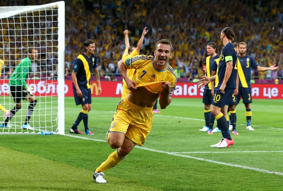A jubilant soccer player in a yellow jersey celebrates after scoring a goal during a match, with teammates and opponents reacting in the background.