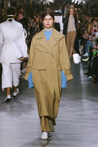 Model wears a long camel trench coat with a blue sweatshirt