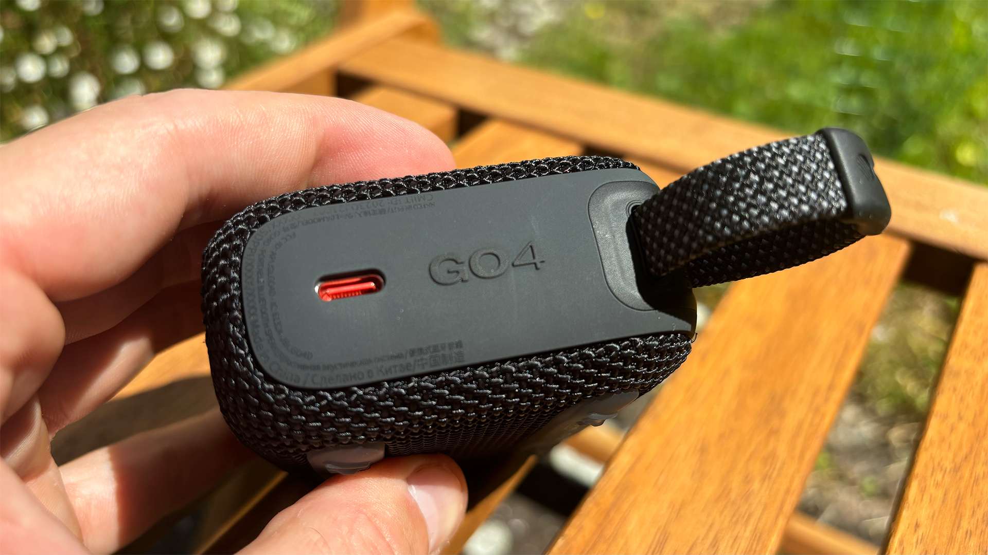 A hand holding a black JBL Go 4 Bluetooth speaker. The carry loop is visible on the right, with the USB-C port on the left.