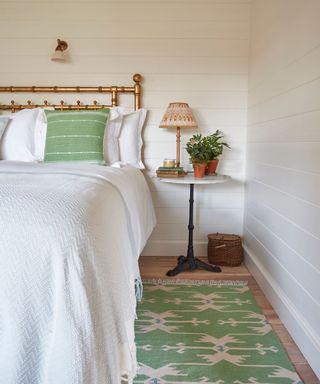 A bedroom with a white bed, pistachio green throw pillows and rug, white walls, and a nightstand with a lamp and plant on it