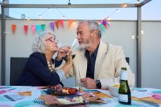 A couple happily celebrates an anniversary at a party, holding champagne flutes for each other to drink.