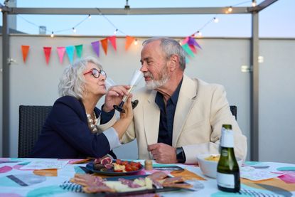 A couple happily celebrates an anniversary at a party, holding champagne flutes for each other to drink. 