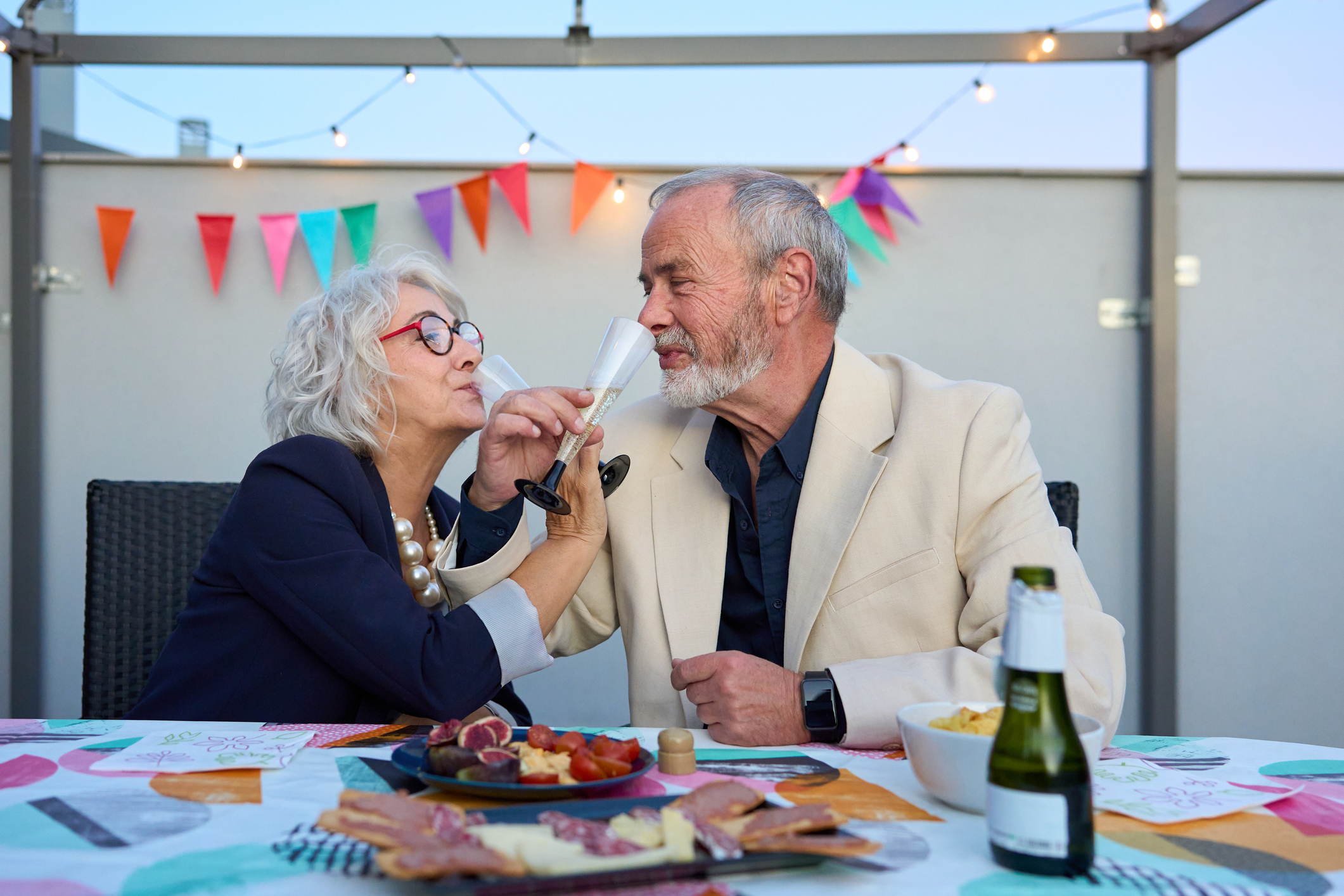 A couple happily celebrates an anniversary at a party, holding champagne flutes for each other to drink. 