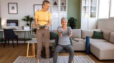 Two women exercise together in a living room. One of the women is sitting in a chair, while the other stands; both are holding dumbbells out in front of them. Behind them is a couch, desk, book shelves and framed pictures. 