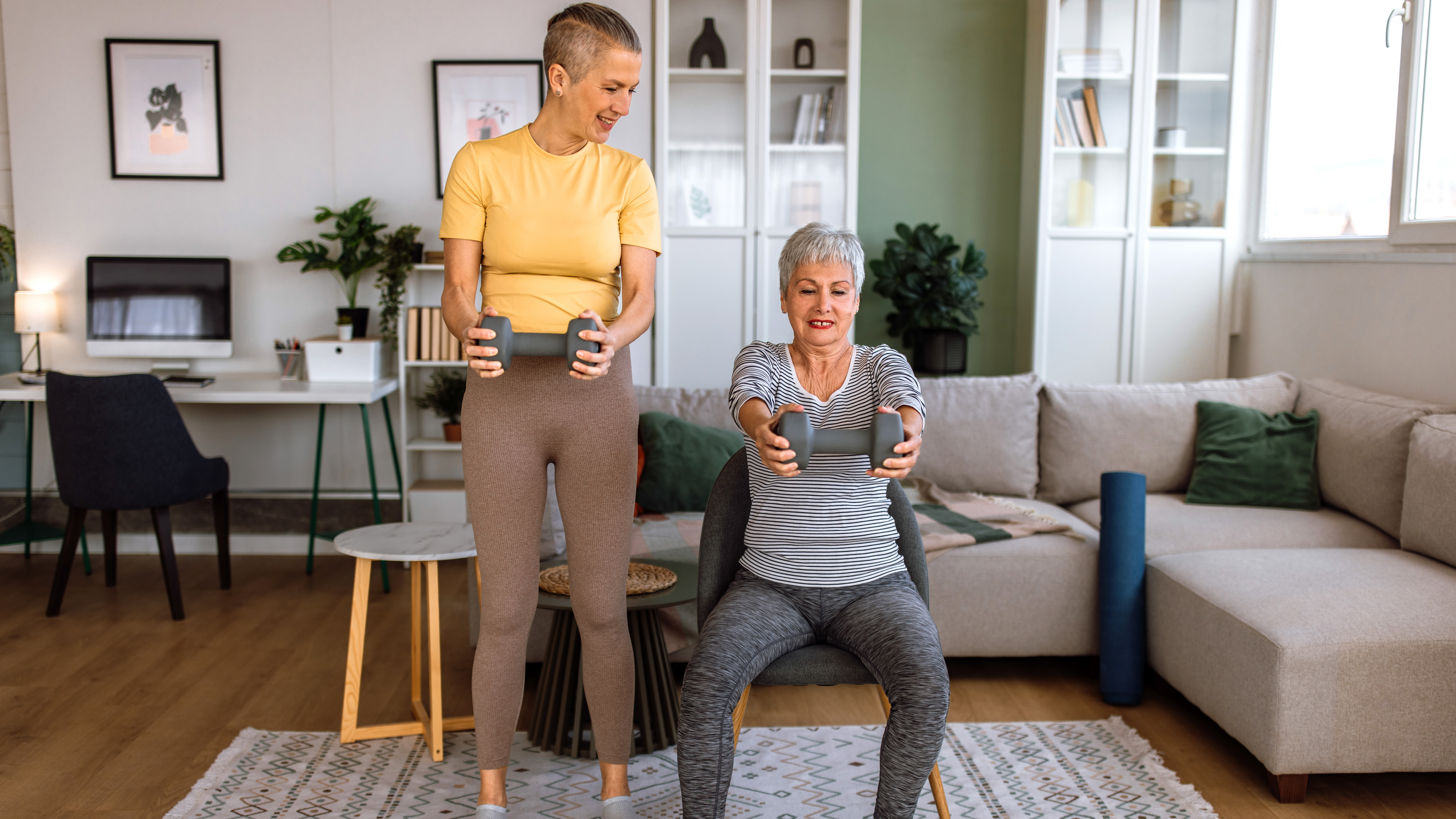 Two women exercise together in a living room. One of the women is sitting in a chair, while the other stands; both are holding dumbbells out in front of them. Behind them is a couch, desk, book shelves and framed pictures. 