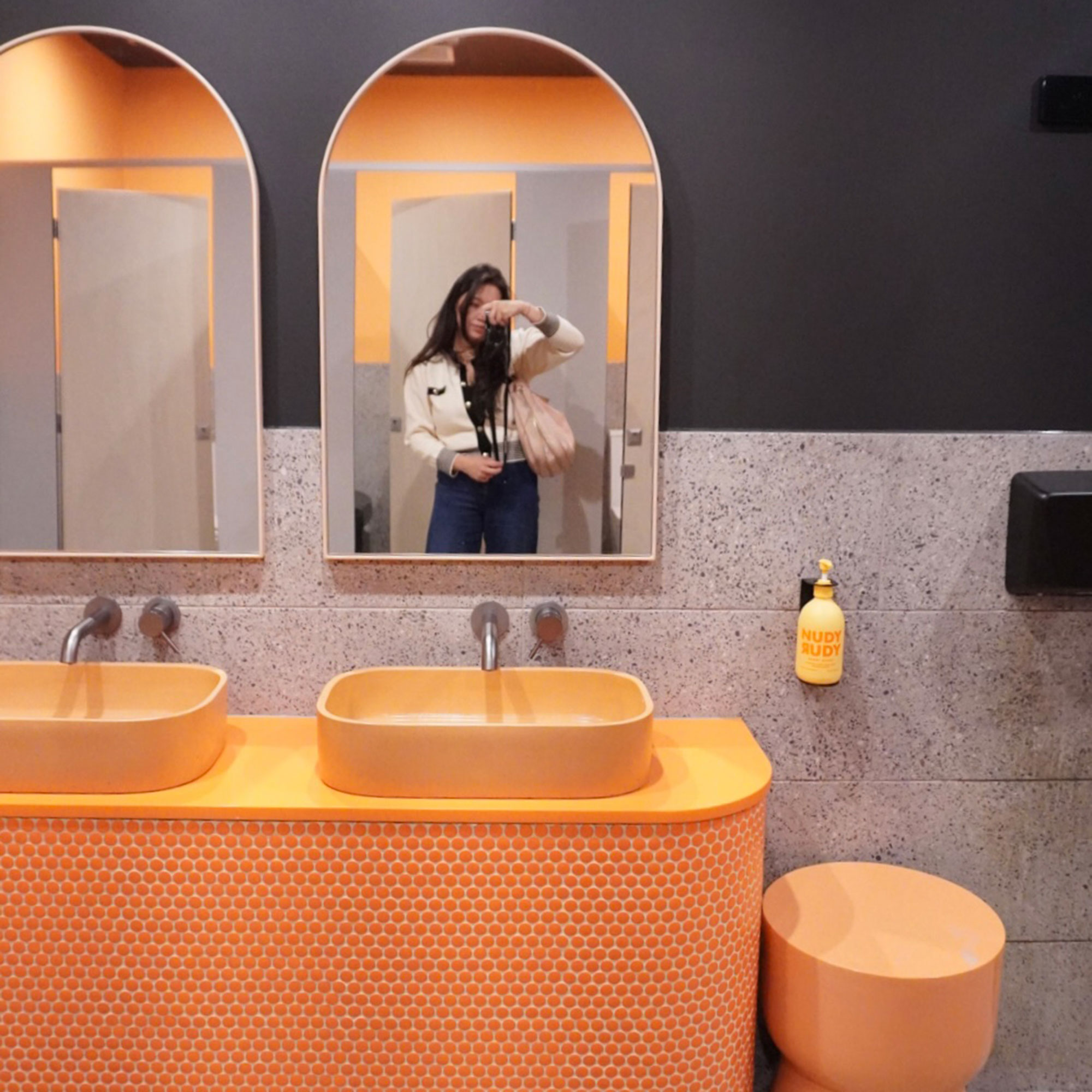 Woman taking selfie in arched bathroom mirror above orange sinks and countertops