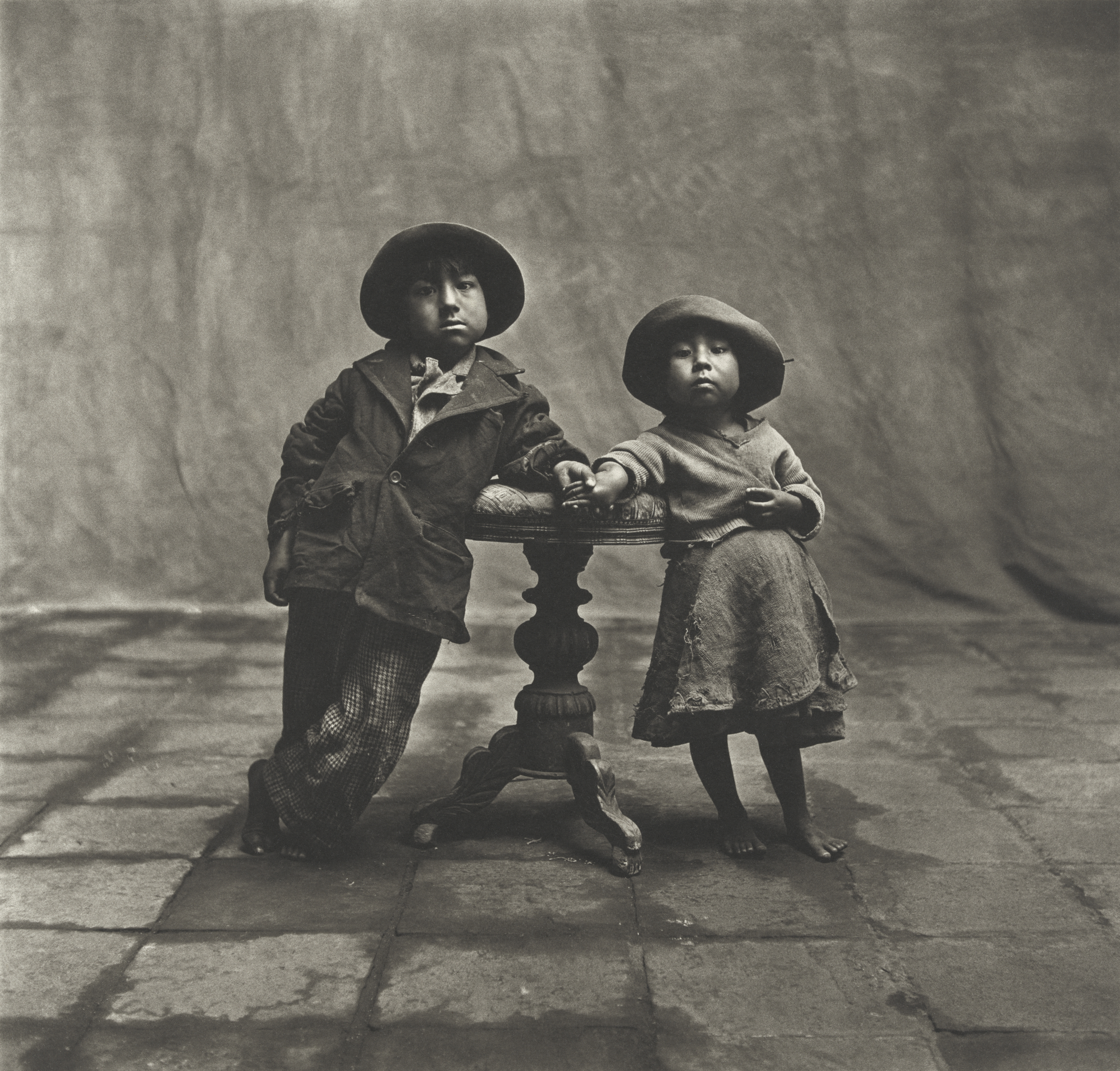 A black-and-white studio portrait by Irving Penn featuring two young children in hats and layered clothing posing barefoot beside a small, ornate wooden table against a draped backdrop.