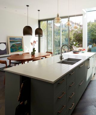 A kitchen island with a white surface and built-in sink and faucet, a dark gray base with brown handles, three glass pendant lights above it, and a wooden dining table next to it