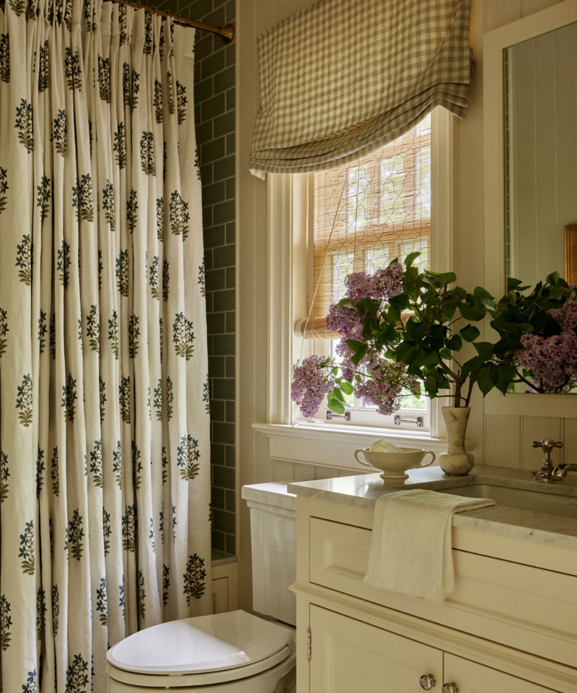 A bathroom interior with a mix of patterns. To the left, a cream shower curtain with a small blue floral motif hangs from a brass rod. A window in the center is covered by a natural wood woven shade and a beige gingham valance. On the marble vanity to the right, a vase of purple lilacs and a small white soap dish sit near the chrome faucet. Sage green subway tiles are visible in the background.
