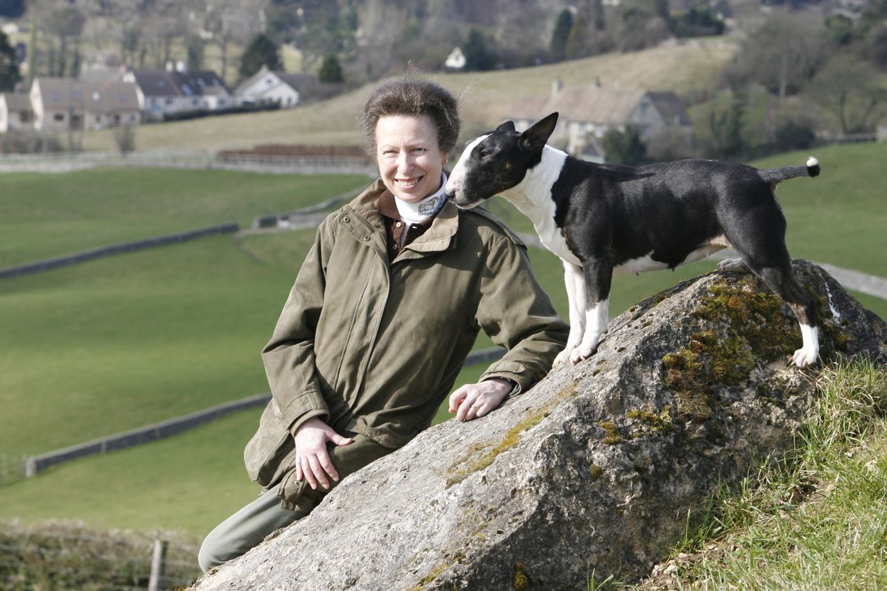 Princess Anne smiles outdoors while her black-and-white Bull Terrier stands on a mossy rock beside her, nuzzling her face