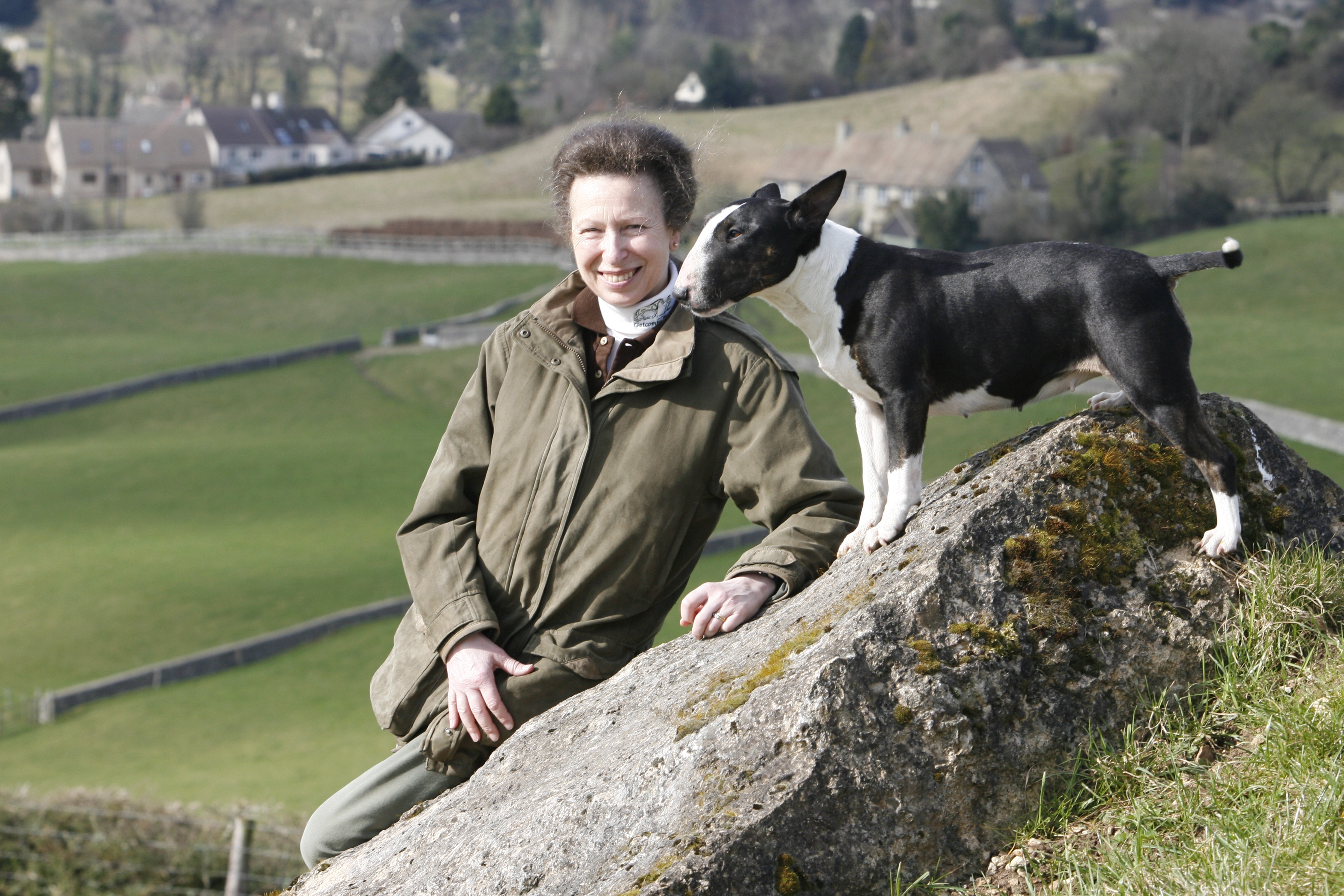 Princess Anne smiles outdoors while her black-and-white Bull Terrier stands on a mossy rock beside her, nuzzling her face
