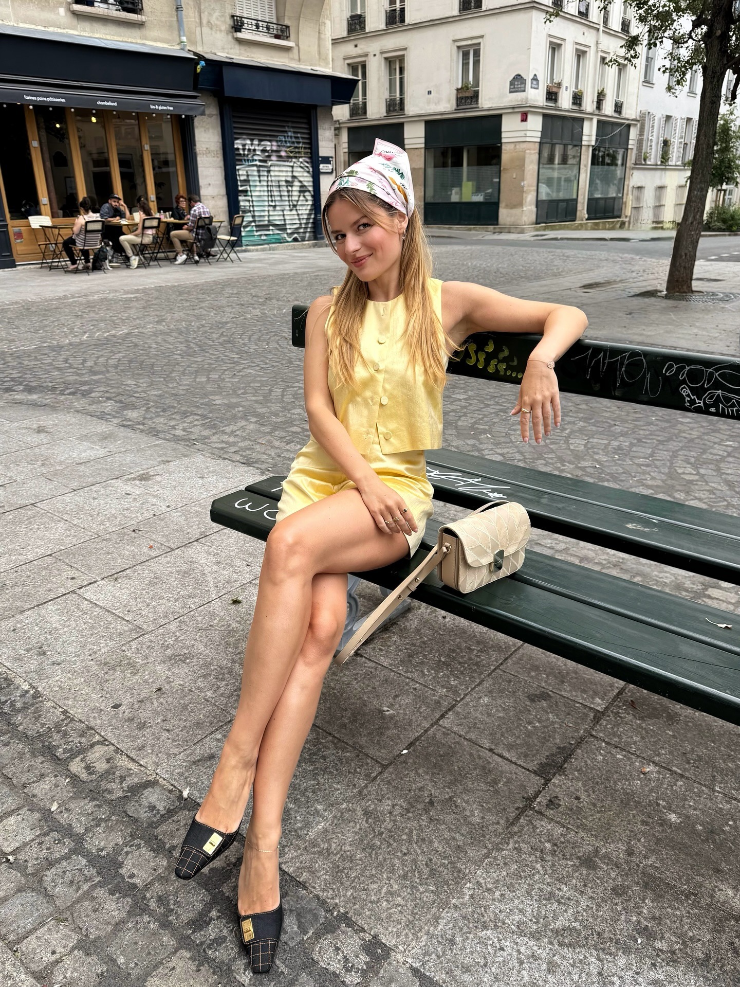 French influencer @sabinasocol sits on a bench outside in Paris wearing a yellow two-piece and black closed-toe heeled mules.