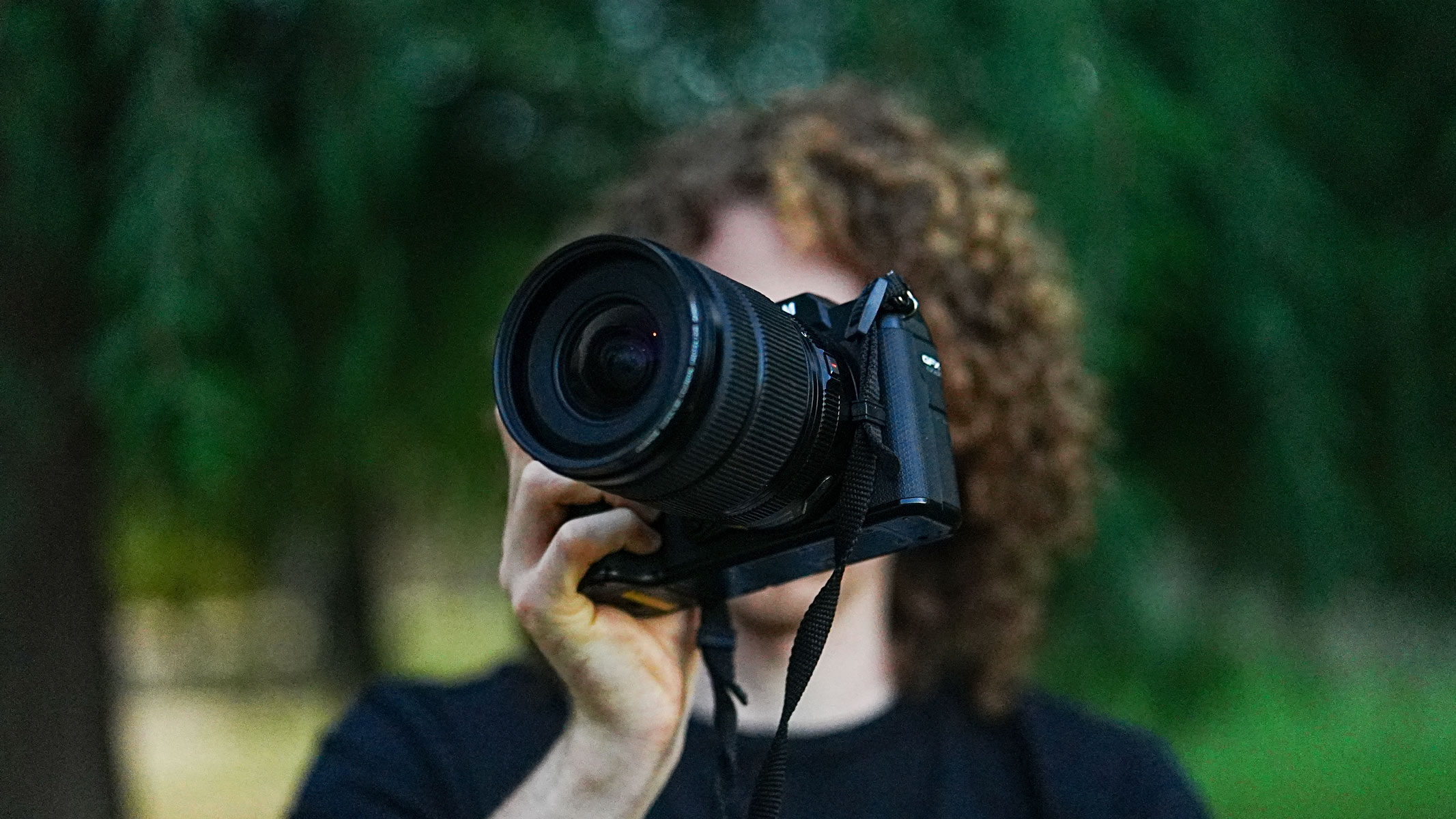 A man with long hair holding the Fujifilm GFX 100S II in front of his face with trees in the background.