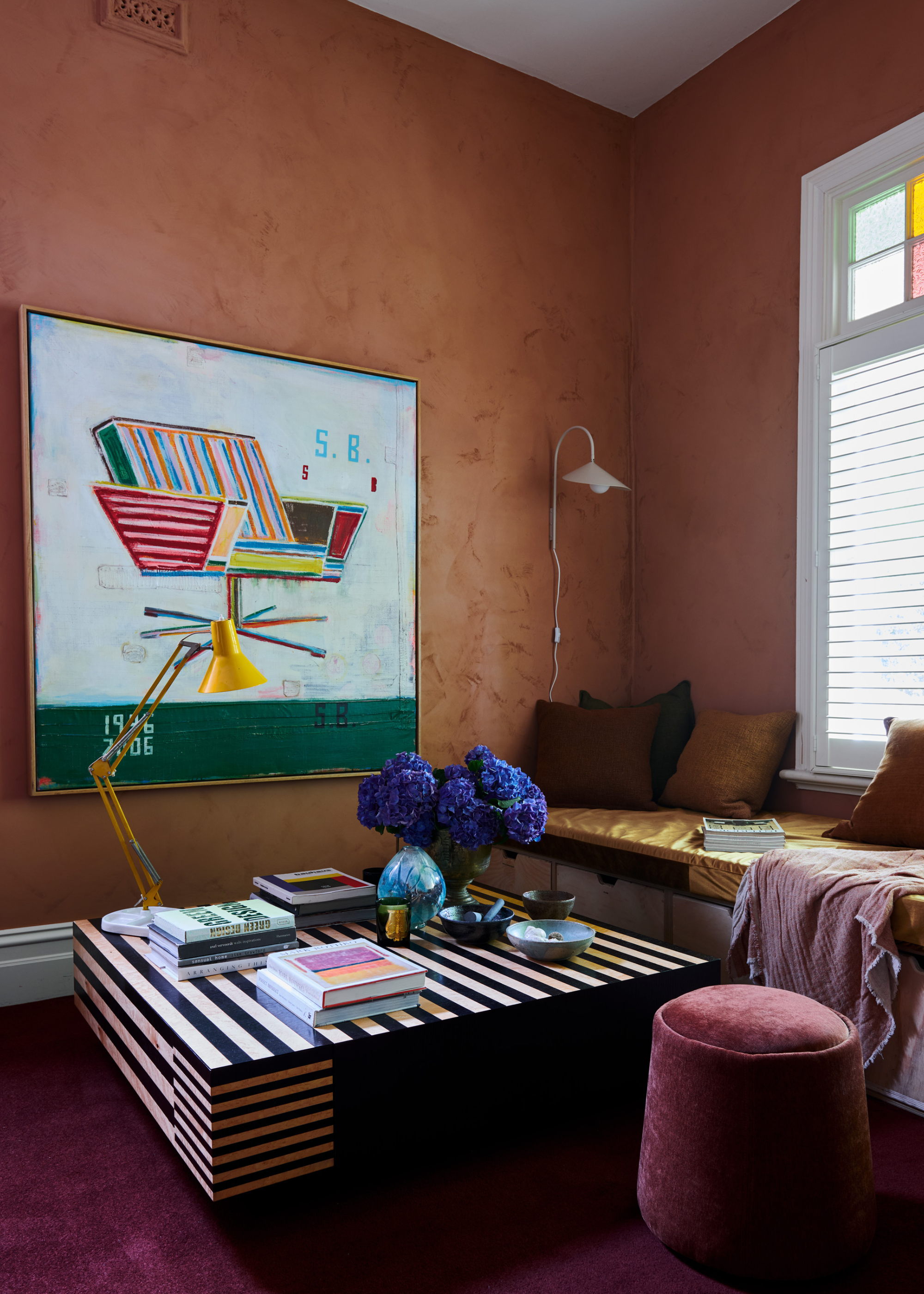 A mustard-brown living nook with a large colorful painting, a beige wall sconce, under-seating storage baskets, a maroon pouffe, a striped coffee table, stacks of books, a yellow table lamp, a vase of purple hydrangeas, and a candle