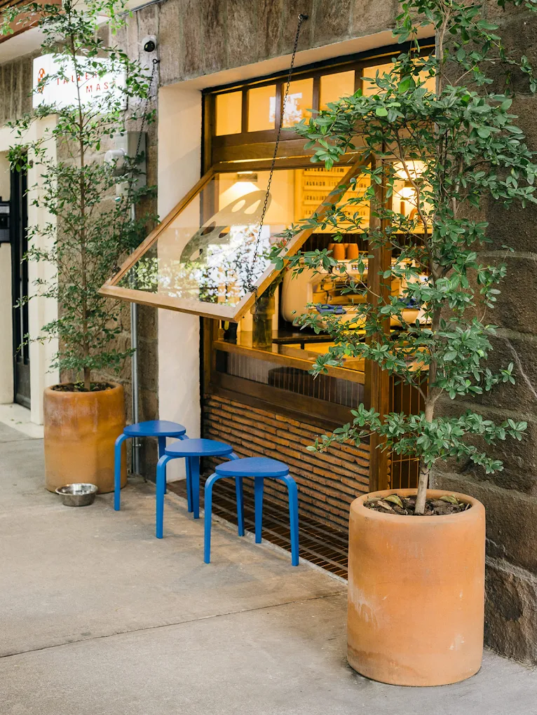 A vibrant, modernist cafe features a golden-lit interior with industrial-style furniture and outdoor punctuated by terracotta planters with thriving plants, minimalist, color-blocked blue stools, brick and cement walls.