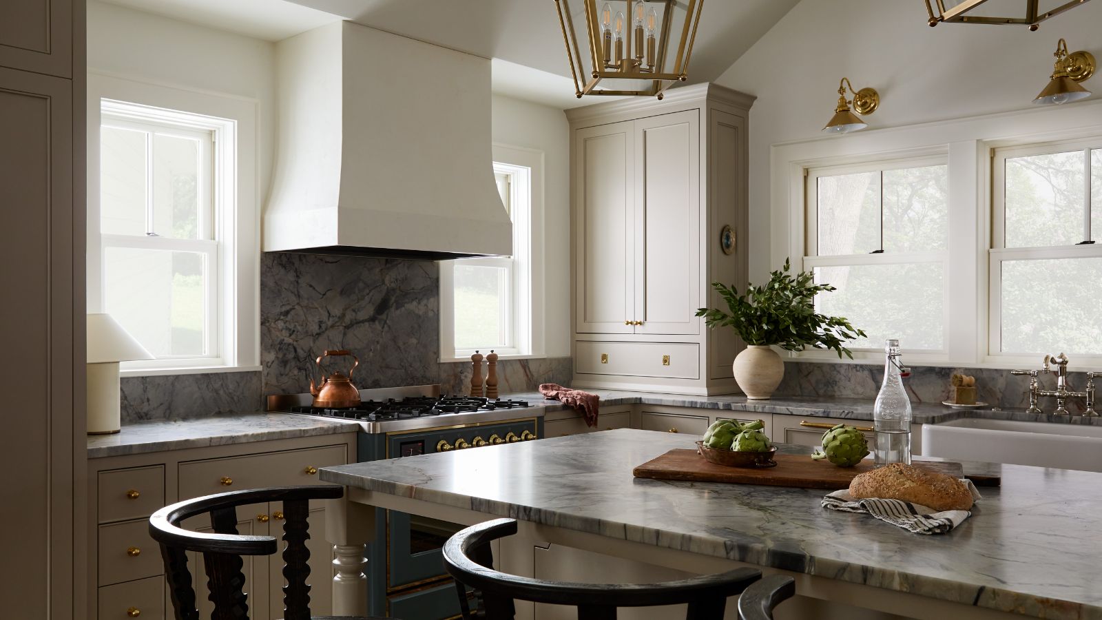 earthy neutral kitchen with large windows out to the lakeside view, a large plaster cooker hood, and blue marble countertops 