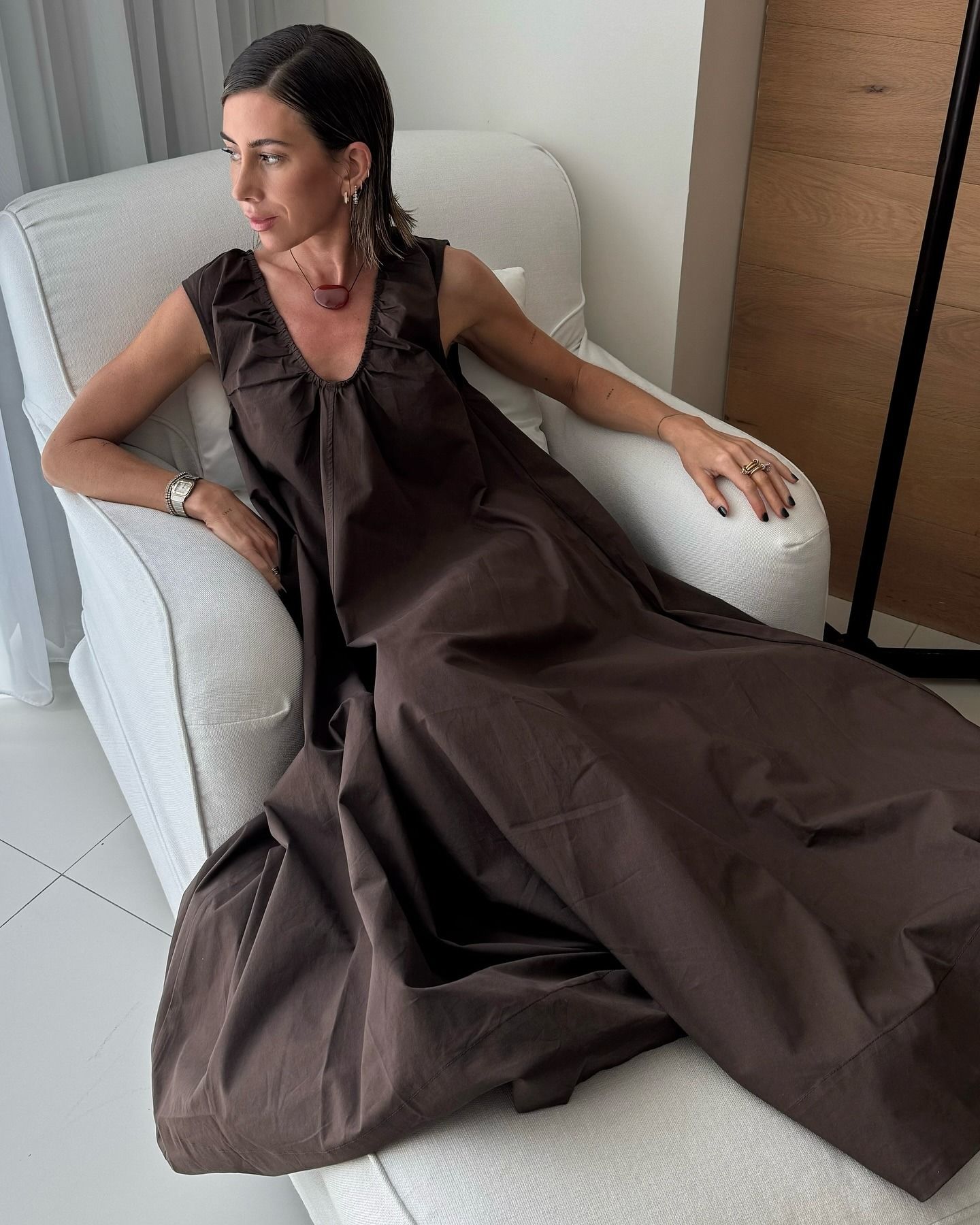 British influencer Hannah Lewis poses in a white linen chair wearing a brown ruched scoop-neck maxi dress and black cord necklace with a red stone.
