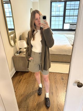 Nikki Chwatt wears Zara bermuda shorts in green with a white tank top, grey cardigan, burgundy loafers, and white socks.