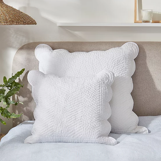 Two stacked white pillows with scalloped edge covers on a bed beside a plant and bedside table. 