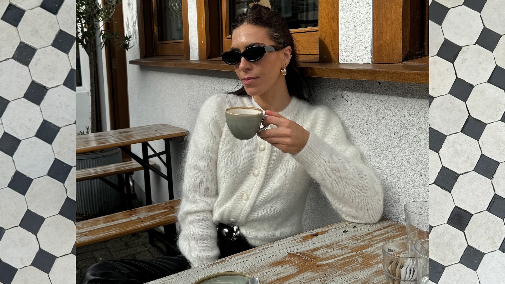 British style influencer Jessica Skye sits at an outdoor cafe in Cornwall drinking a cup of coffee wearing black oval sunglasses, a fuzzy off white cardigan, a studded belt, and leather pants.