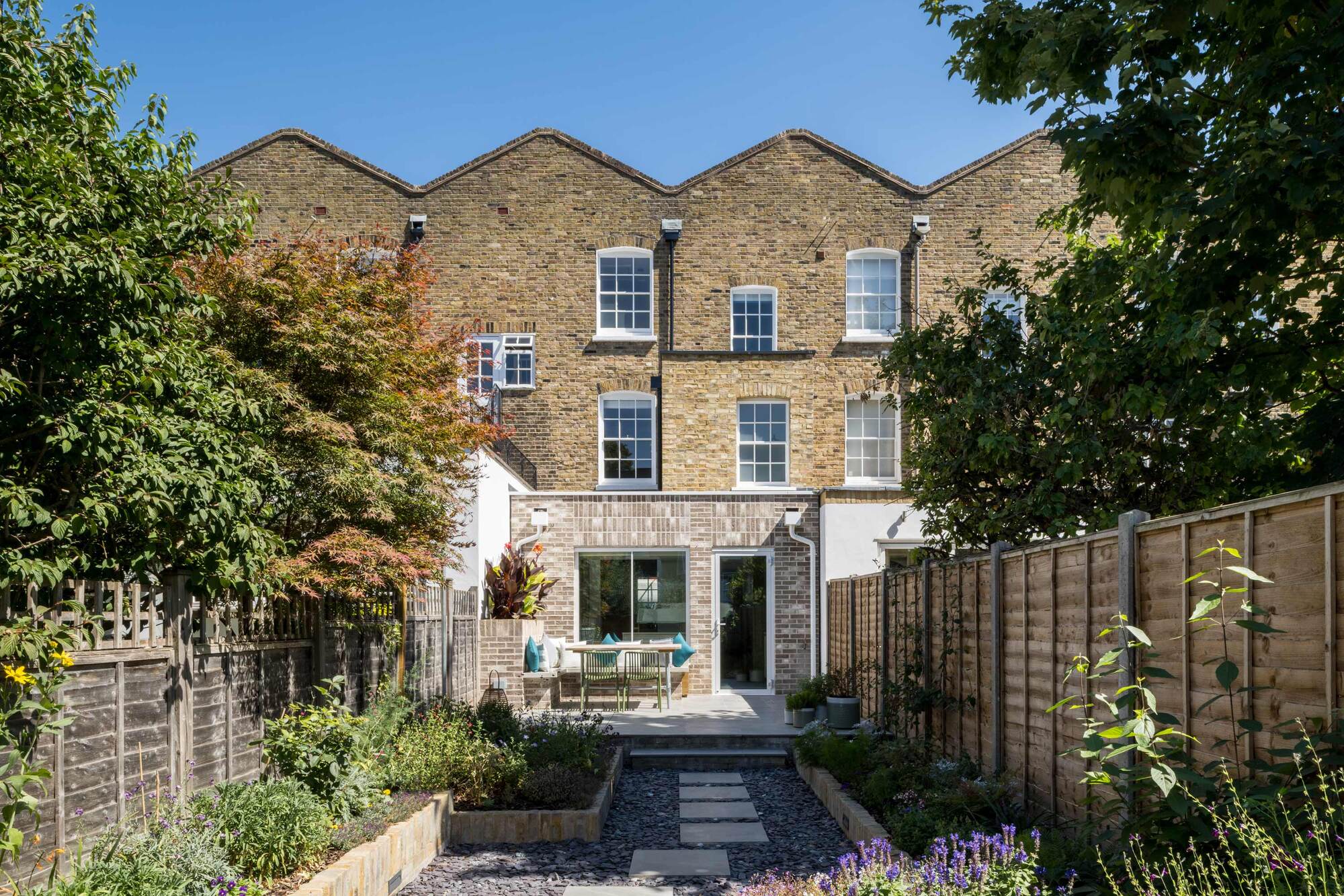a narrow terrace garden with L-shaped paved garden path and a deck with table and chairs in the sun, and planter boxes running along the fenceline